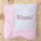 May include: White and pink baby quilt with the name "Hazel" embroidered in floral letters. The quilt features a detailed quilted pattern and a pink border. A soft and cozy baby blanket.