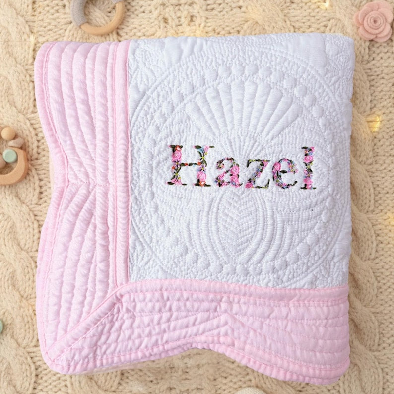 May include: White and pink baby quilt with the name "Hazel" embroidered in floral letters. The quilt features a detailed quilted pattern and a pink border. A soft and cozy baby blanket.