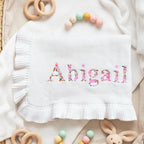 May include: White baby blanket with the name "Abigail" embroidered in pink floral letters. The blanket has a ruffled edge and is surrounded by wooden toys and a plush bunny.