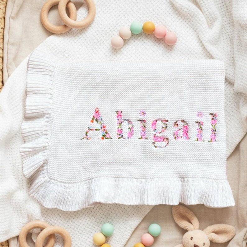 May include: White baby blanket with the name "Abigail" embroidered in pink floral letters. The blanket has a ruffled edge and is surrounded by wooden toys and a plush bunny.