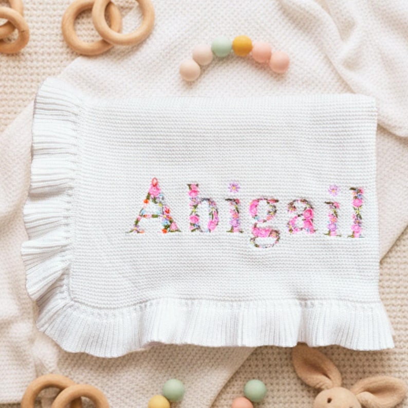 May include: A white baby blanket with the name "Abigail" embroidered in a floral pattern. The blanket has a ruffled edge and is surrounded by wooden toys and a teething ring.