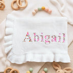 May include: A white baby blanket with the name "Abigail" embroidered in a floral pattern. The blanket has a ruffled edge and is surrounded by wooden toys and a teething ring.