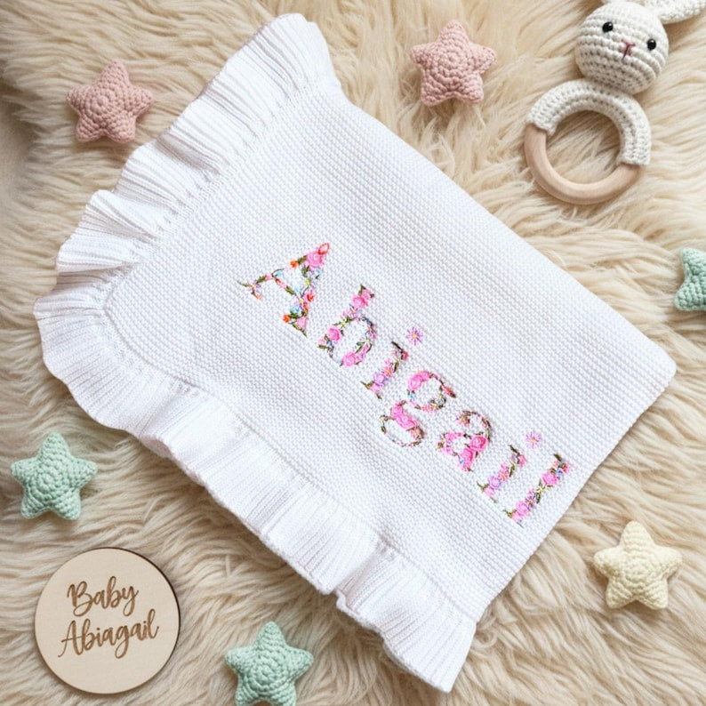 May include: White baby blanket with a ruffled edge and the name "Abigail" embroidered in floral letters. The blanket is surrounded by star-shaped toys and a wooden disc that says "Baby Abigail".