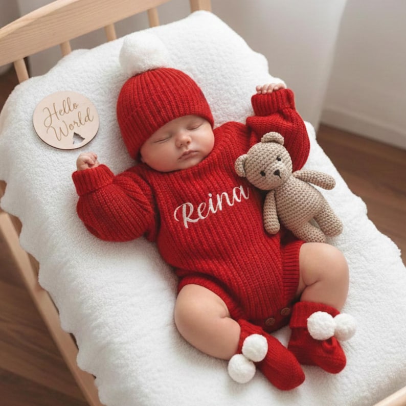 May include: A sleeping baby wearing a red knit outfit, including a hat with a white pom-pom, a sweater with the name "Reina", and matching booties with white pom-poms. A small, beige teddy bear and a wooden disc with "Hello World" are also