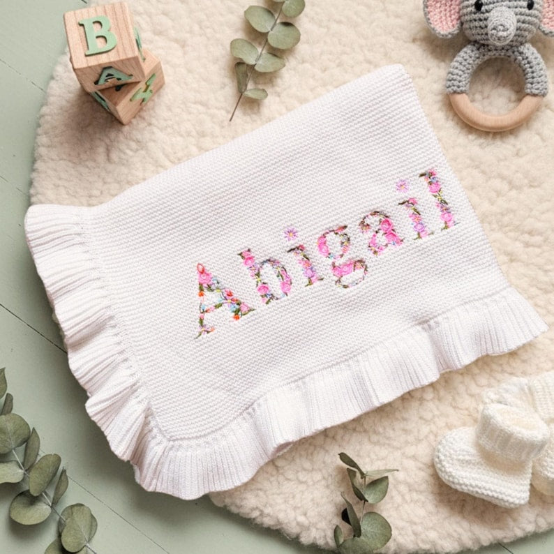 May include: White baby blanket with the name "Abigail" embroidered in a floral pattern. The blanket has a ruffled edge and is surrounded by baby accessories, including a wooden block and a crochet elephant toy.
