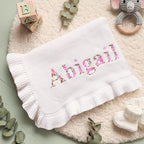 May include: White baby blanket with the name "Abigail" embroidered in a floral pattern. The blanket has a ruffled edge and is surrounded by baby accessories, including a wooden block and a crochet elephant toy.