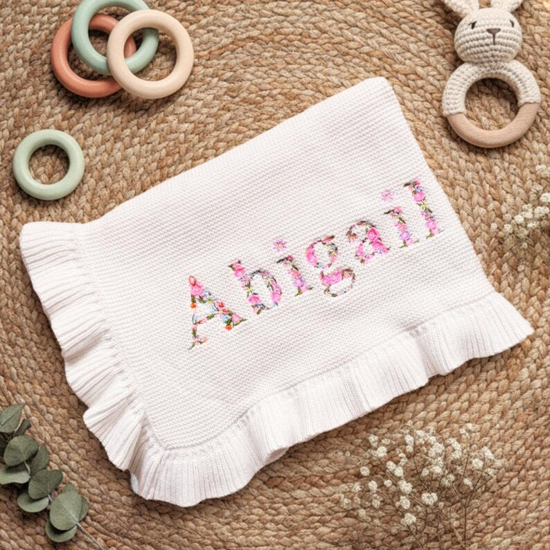 May include: A white knitted baby blanket with a ruffled edge. The name "Abigail" is embroidered in floral print across the center. The blanket is surrounded by wooden baby toys, including a bunny rattle and colorful rings.