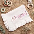 May include: A white knitted baby blanket with a ruffled edge. The name "Abigail" is embroidered in floral print across the center. The blanket is surrounded by wooden baby toys, including a bunny rattle and colorful rings.