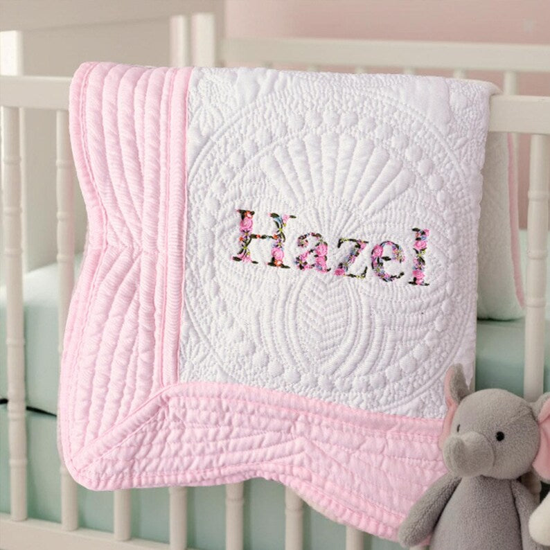 May include: A white baby quilt with pink trim and the name "Hazel" embroidered in floral fabric. The quilt features intricate quilted patterns and is draped over a white crib. A gray elephant plush toy is in the foreground.