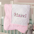 May include: A white baby quilt with pink trim and the name "Hazel" embroidered in floral fabric. The quilt features intricate quilted patterns and is draped over a white crib. A gray elephant plush toy is in the foreground.