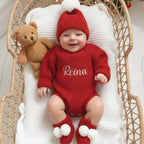 May include: A baby wearing a red knit outfit, including a hat with a white pom-pom, a long-sleeved bodysuit with the name "Reina" embroidered, and matching booties with white pom-poms. A brown teddy bear is in the wicker basket.