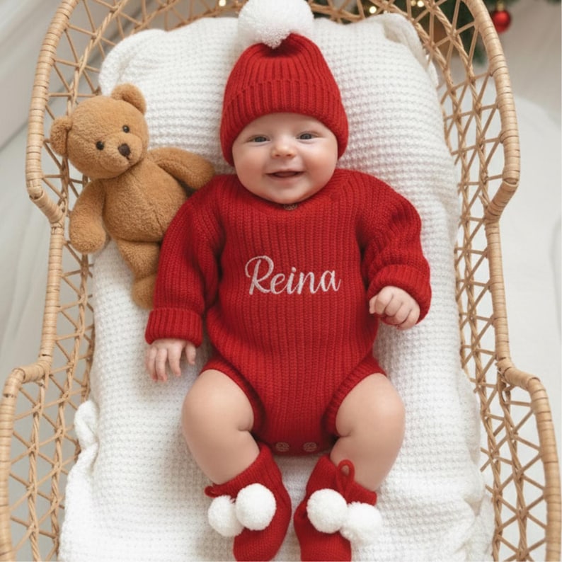 May include: A baby wearing a red knit outfit, including a hat with a white pom-pom, a long-sleeved bodysuit with the name "Reina" embroidered, and matching booties with white pom-poms. A brown teddy bear is in the wicker basket.
