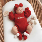 May include: A sleeping baby wearing a red knit outfit, including a bodysuit with the name "Reina" embroidered in white, a headband with a bow, and matching booties with white pom-poms. A small, beige teddy bear and a wooden disc that says 