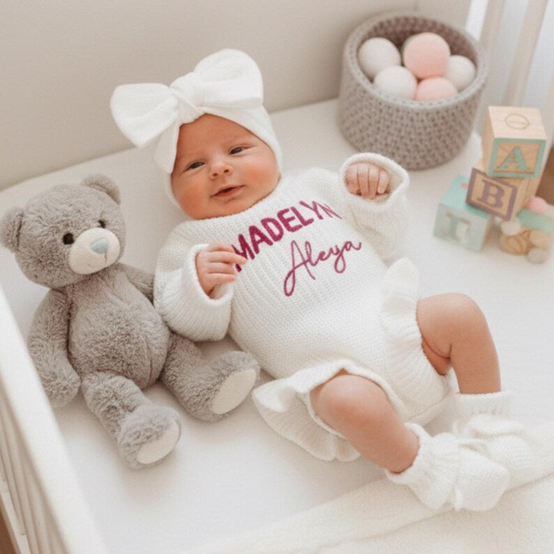 May include: A newborn baby wearing a white knit romper with the name "Madelyn Aleya" in purple script. The baby is also wearing a matching bow headband and booties. A gray teddy bear sits nearby. A basket of balls and wooden blocks are in 