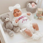 May include: A baby wearing a white knit romper with the name "Madelyn Aleya" in pink script, a matching bow headband, and ruffled leg warmers. A gray teddy bear sits beside the baby in a crib. Wooden blocks and a basket of decorative balls