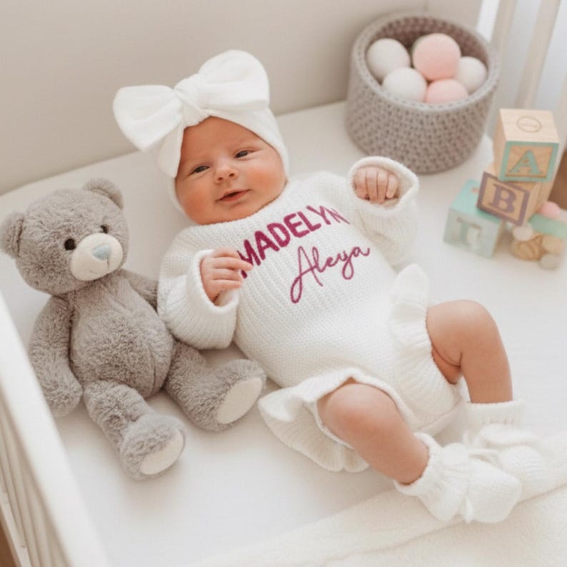 May include: A baby wearing a white knit romper with the name "Madelyn Aleya" in pink script, a matching bow headband, and ruffled leg warmers. A gray teddy bear sits beside the baby in a crib. Wooden blocks and a basket of decorative balls