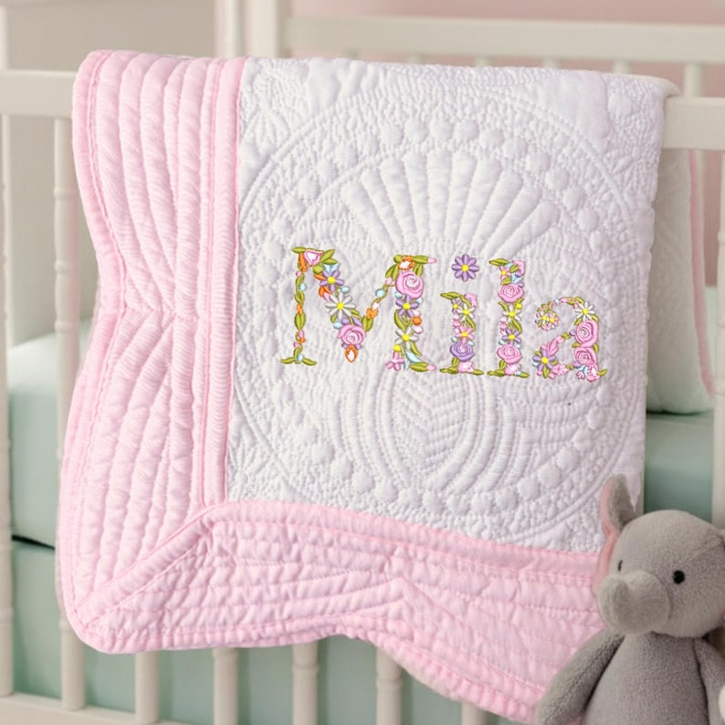 May include: A white and pink baby quilt with the name "Mila" embroidered in colorful floral letters. The quilt has a detailed quilted pattern and a pink border. A gray stuffed elephant is in the lower right corner.
