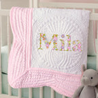 May include: A white and pink baby quilt with the name "Mila" embroidered in colorful floral letters. The quilt has a detailed quilted pattern and a pink border. A gray stuffed elephant is in the lower right corner.
