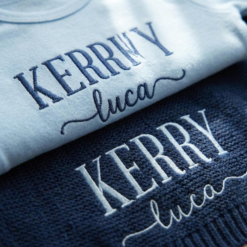 May include: A light blue baby onesie and a navy blue knit blanket, both embroidered with the name "Kerry" and "luca" in a cursive font. The onesie is made of soft material, and the blanket has a textured knit pattern.