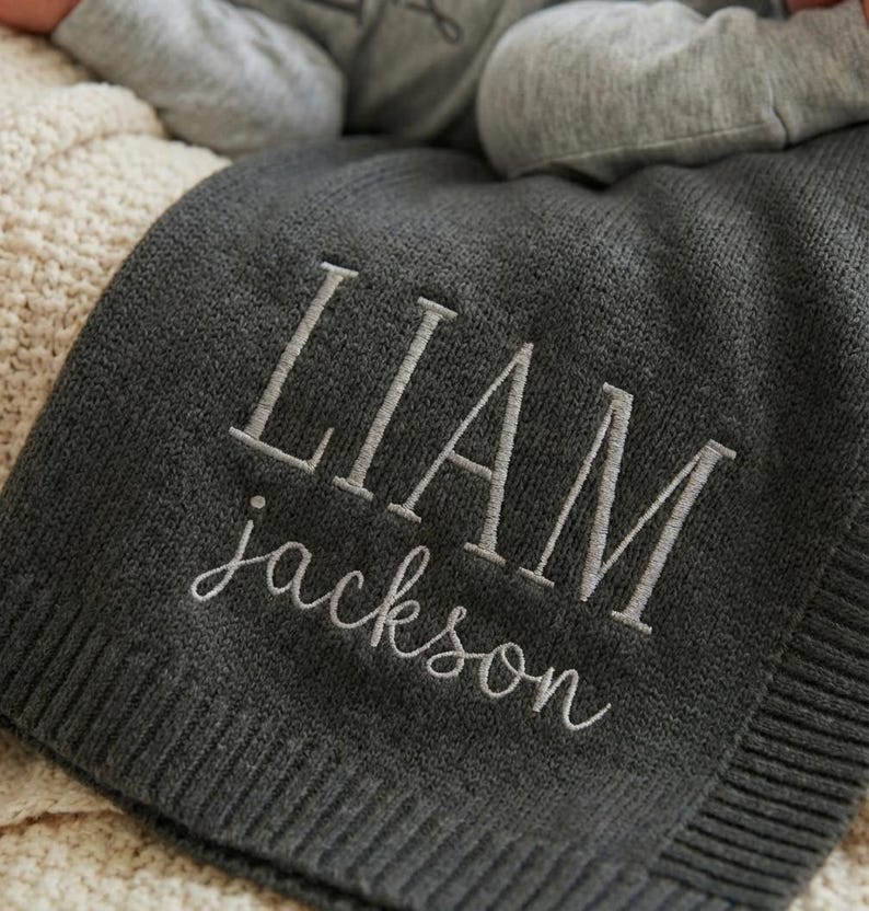 May include: A dark gray knit baby blanket with the name "LIAM" and the last name "jackson" embroidered in a lighter gray thread. The blanket is folded and appears soft and cozy.
