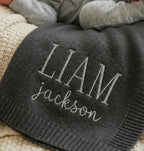 May include: A dark gray knit baby blanket with the name "LIAM" and the last name "jackson" embroidered in a lighter gray thread. The blanket is folded and appears soft and cozy.