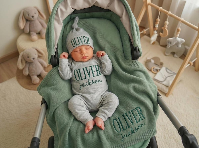 May include: A newborn baby wearing a gray outfit and hat, both embroidered with the name "OLIVER" in green, is nestled in a green stroller. A matching blanket with the name "OLIVER Jackson" is draped over the baby. Plush bunny toys are nea