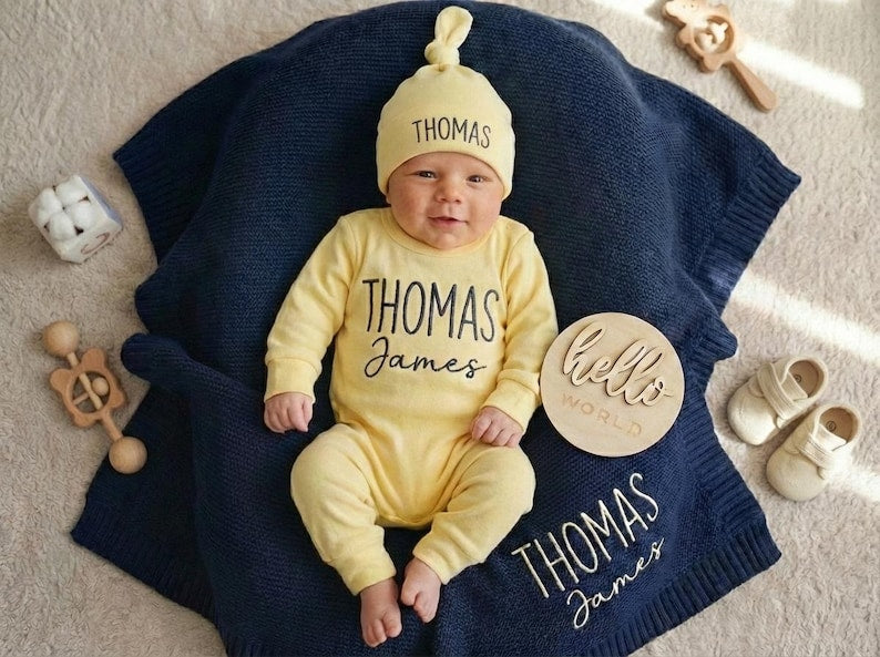 May include: A baby wearing a yellow onesie and hat with the name "Thomas" printed on them, lying on a navy blue blanket. A wooden sign says "hello world". There are baby shoes and toys nearby.