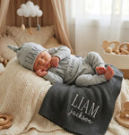 May include: A newborn baby wearing a gray hat, long-sleeved onesie, and blanket, all embroidered with the name "LIAM Jackson" in white. The baby is lying on a cream-colored knitted blanket, with a gray blanket underneath. A cloud mobile ha