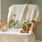 May include: Cream-colored knitted baby blanket with the name "Oliver" embroidered in green script. The blanket is decorated with embroidered bees and daisies. Two small brown teddy bears sit on the blanket, draped over a wooden rocking cha