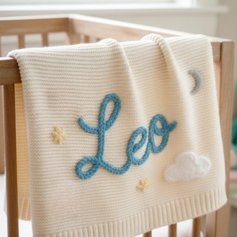 May include: Cream-colored knitted baby blanket with the name "Leo" in blue script. The blanket features a cloud and star embellishments, and a gray crescent moon. The blanket is draped over a wooden crib.