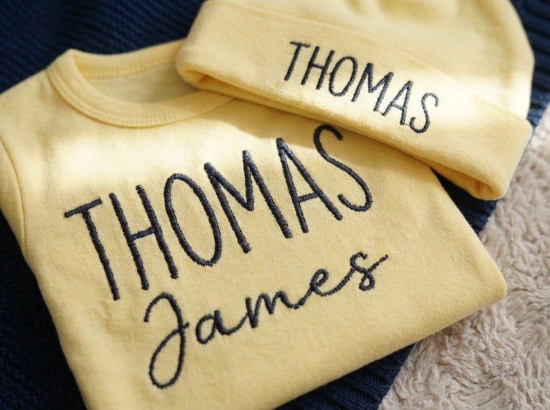 May include: A light yellow baby outfit set, including a long-sleeved shirt and a beanie. The name "THOMAS" is embroidered on the beanie and shirt. The shirt also has the name "James" embroidered below "THOMAS".
