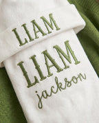 May include: A white baby beanie and blanket set with the name "LIAM" embroidered in green. The blanket also has the name "Jackson" embroidered in green. The set is displayed on a green background.