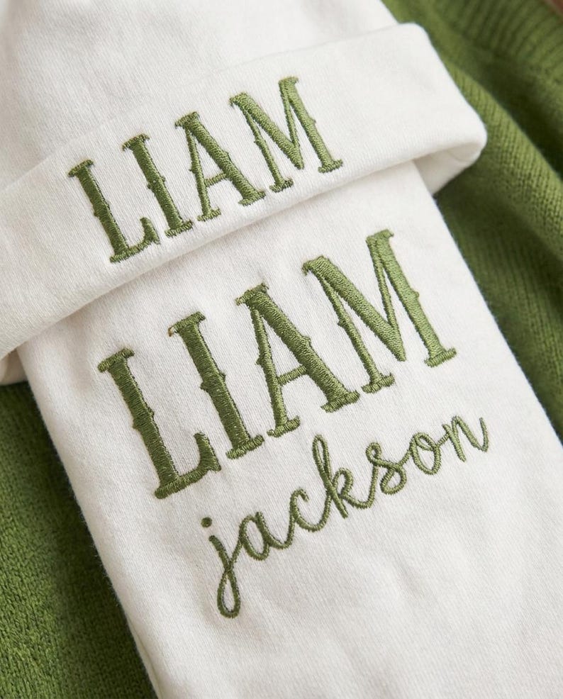 May include: A white baby beanie and blanket set with the name "LIAM" embroidered in green. The blanket also has the name "Jackson" embroidered in green. The set is displayed on a green background.