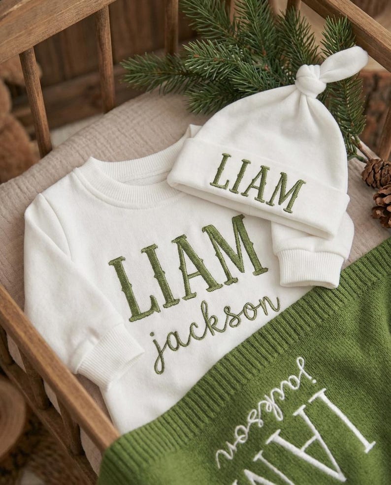 May include: A white baby outfit set with the name "LIAM" embroidered in green on a sweatshirt and hat. A green blanket with the name "jackson" embroidered in white is also included. The set is displayed in a wooden crib.