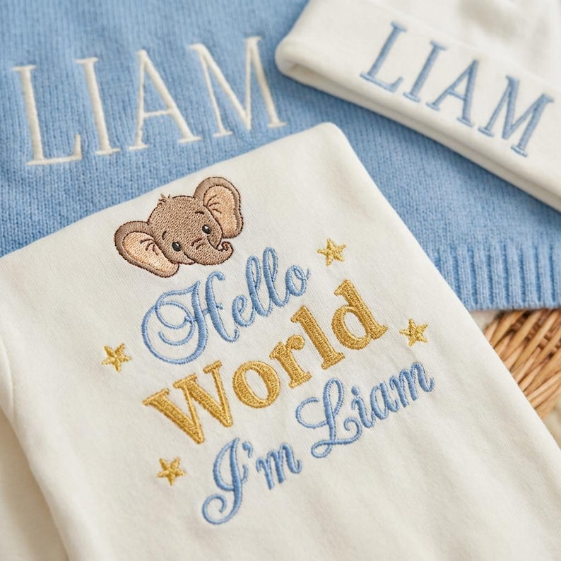 May include: A baby gift set featuring a white onesie with an embroidered elephant and the text "Hello World I'm Liam". A light blue knit blanket and a white knit hat, both embroidered with the name "Liam", complete the set. The set is desi