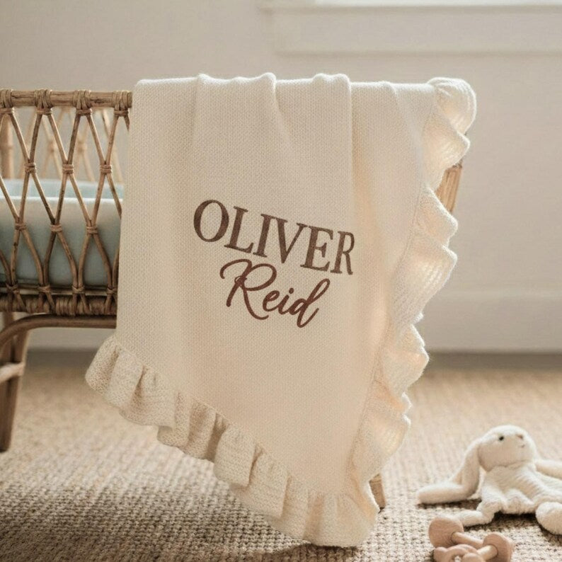 May include: Cream-colored baby blanket with a ruffled edge, draped over a crib. The blanket features the embroidered name "OLIVER Reid" in a brown script font. A plush bunny toy and a wooden rattle are in the foreground.