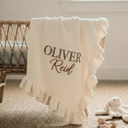 May include: Cream-colored baby blanket with a ruffled edge, draped over a crib. The blanket features the embroidered name "OLIVER Reid" in a brown script font. A plush bunny toy and a wooden rattle are in the foreground.
