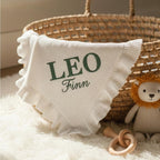 May include: A white baby blanket with a ruffled edge, embroidered with the name "LEO" and "Finn" in green. The blanket is in a woven basket with a stuffed lion toy and a wooden rattle.