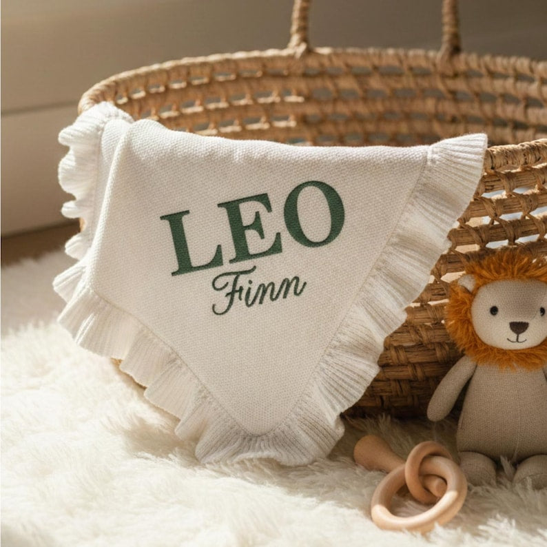 May include: A white baby blanket with a ruffled edge, embroidered with the name "LEO" and "Finn" in green. The blanket is in a woven basket with a stuffed lion toy and a wooden rattle.