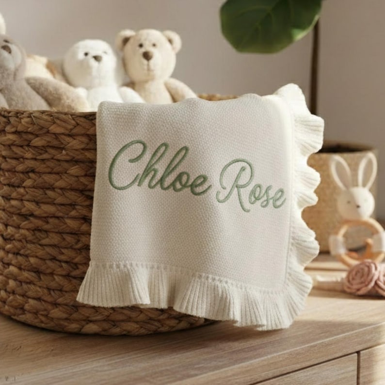 May include: A cream-colored baby blanket with a ruffled edge and the embroidered name "Chloe Rose" in green. The blanket is in a woven basket with stuffed animals.