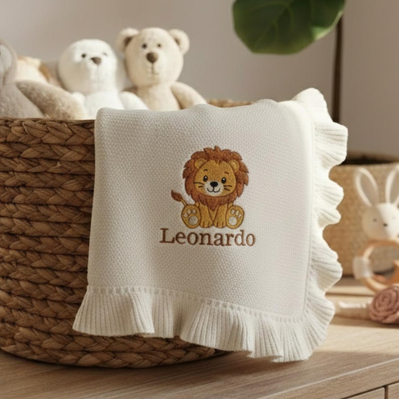 May include: Cream-colored baby blanket with a ruffled edge. The blanket features an embroidered lion design in brown and tan, with the name "Leonardo" below. The blanket is folded and placed in a woven basket with stuffed animals.
