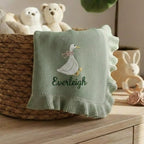 May include: A light green baby blanket with a ruffled edge. The blanket features an embroidered white goose with a pink bow and the name "Everleigh" in green script. The blanket is displayed in a woven basket with stuffed animals.
