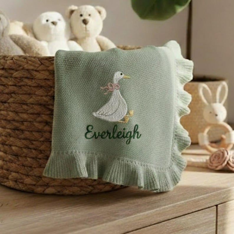 May include: A light green baby blanket with a ruffled edge. The blanket features an embroidered white goose with a pink bow and the name "Everleigh" in green script. The blanket is displayed in a woven basket with stuffed animals.