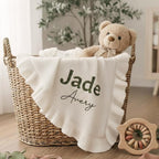 May include: A woven basket holds a white baby blanket with a ruffled edge, embroidered with the names "Jade" and "Avery" in green and black. A teddy bear with a bow sits in the basket. A wooden toy with colorful rings is next to the basket