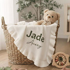 May include: A woven basket holds a white baby blanket with a ruffled edge and the names "Jade Avery" embroidered in green and black. A teddy bear with a bow sits in the basket, along with a wooden toy.