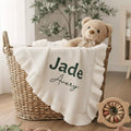 May include: A woven basket holds a white baby blanket with a ruffled edge, embroidered with the names "Jade Avery" in green. A teddy bear and baby clothes are inside. A wooden toy with colorful rings sits nearby.