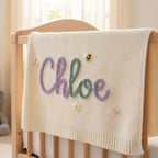 May include: Cream-colored baby blanket with the name "Chloe" in purple and teal script. The blanket is decorated with a small bee, a daisy, and two butterflies. The blanket is draped over a wooden crib.