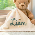 May include: Cream-colored baby blanket with the name "Liam" embroidered in teal script. The blanket is decorated with a small bee, flowers, and a butterfly. A teddy bear sits behind the blanket.