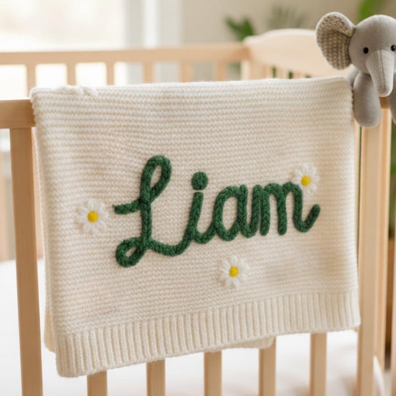 May include: Cream-colored knitted baby blanket with the name "Liam" in green script. The blanket is decorated with three white daisies with yellow centers. The blanket is draped over a wooden crib.