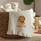 May include: Cream-colored baby blanket with a ruffled edge. The blanket features an embroidered lion wearing a crown and a red scarf, with the text "Dedric King" below. The blanket is folded over a wicker basket, with stuffed animals in th